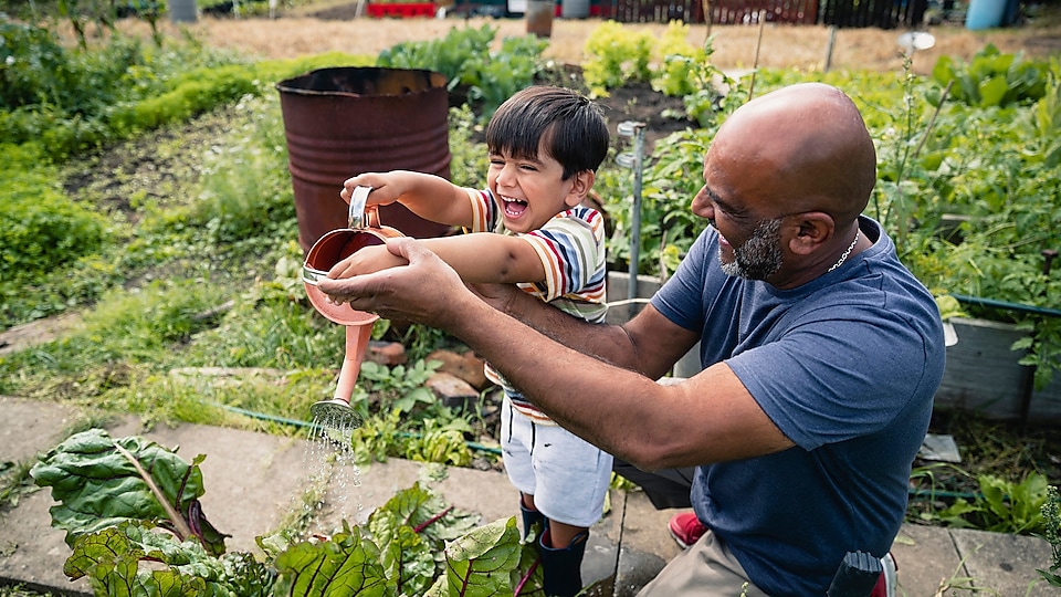 Man helping a smiling child water plants in a garden