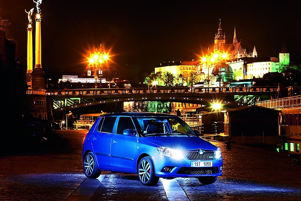 Blue car parked on a Prague road with a bridge and castle in the backdrop, at night