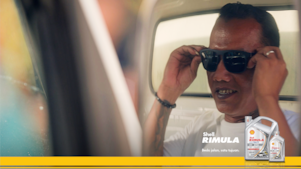 Seorang pengemudi truk mengenakan kacamata hitam sambil tersenyum, dengan teks promosi Shell Rimula LD4 Extra dan kemasan produk di sisi kanan.