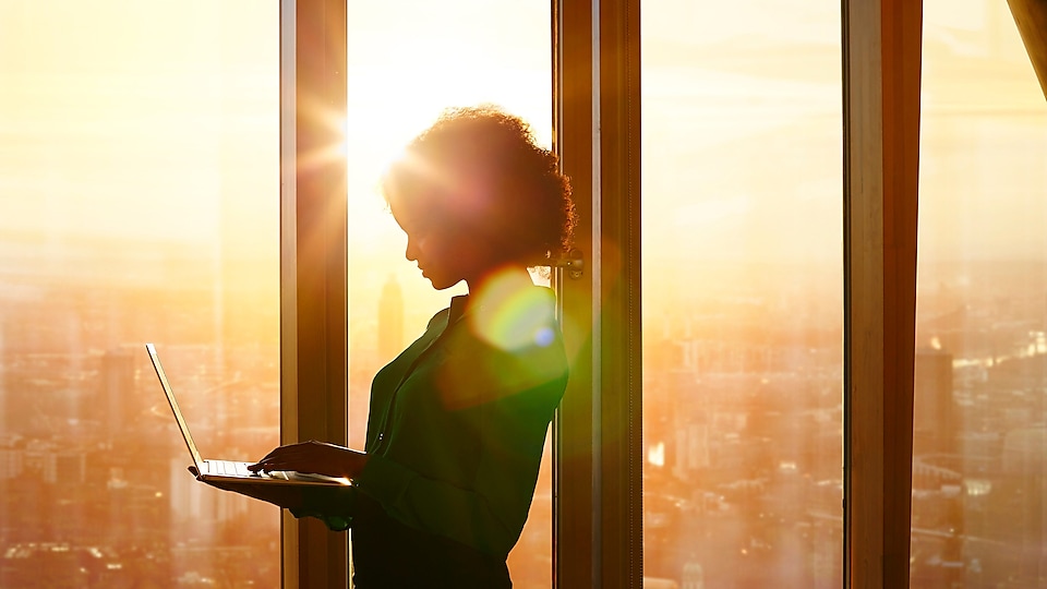 Seorang wanita berdiri membawa laptop dengan latar belakang matahari terbenam di balik jendela di sebuah gedung