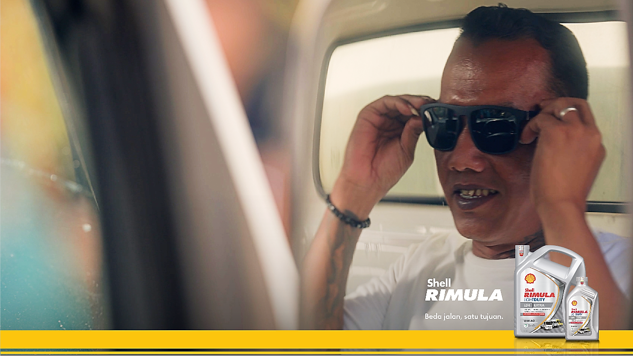 Seorang pengemudi truk mengenakan kacamata hitam sambil tersenyum, dengan teks promosi Shell Rimula LD4 Extra dan kemasan produk di sisi kanan.
