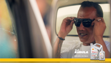 Seorang pengemudi truk mengenakan kacamata hitam sambil tersenyum, dengan teks promosi Shell Rimula LD4 Extra dan kemasan produk di sisi kanan.